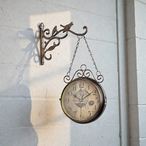 May include: A double-sided, antique-style clock with a bird and cage design on the face. The clock hangs from a decorative metal bracket with a bird and leaf accents. The clock face has black numerals and hands.