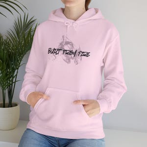 Peut inclure: Un sweat à capuche rose clair avec une poche avant et une capuche à cordon. Le texte "BUILT FROM FIRE" est imprimé en noir sur la poitrine, avec un motif de fumée derrière le texte. Le sweat à capuche est en matière douce.