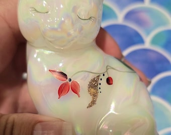 May include: Iridescent white ceramic cat figurine with closed eyes and floral accents. The cat has a pearlescent finish and is decorated with red and gold painted details. A decorative item.