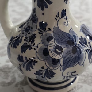 Könnte beinhalten: Kleine, weiße Keramikkrug mit Henkel und Ausguss. Der Krug ist mit blauen Blumenmustern verziert. Der Sockel hat ein kreisförmiges Design mit blauen Streifen. Zum Servieren von Flüssigkeiten.