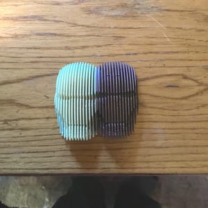 May include: Two abstract, geometric sculptures, each composed of vertical, parallel lines. One is light green, the other is dark purple. The sculptures are placed side-by-side on a wooden surface.