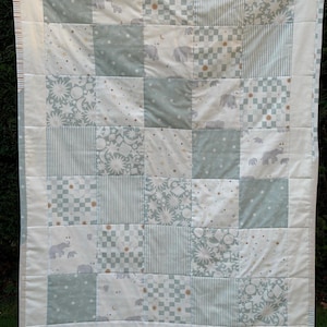 May include: A patchwork baby quilt in white, pale blue, and grey. The quilt features squares with elephant motifs, stripes, stars, and checkered patterns. The quilt is bordered with white fabric. The quilt is likely made of cotton.