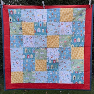 May include: A patchwork quilt with a red border and blue backing, hanging on a washing line. The quilt is made up of squares with beach hut, boat, and yellow polka dot patterns. The quilt is made of various fabrics.