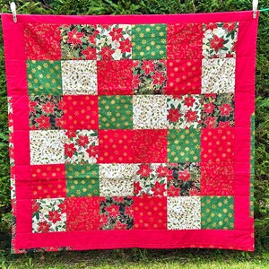May include: A Christmas-themed patchwork quilt with a red border. The quilt is made up of squares in red, green, and white, decorated with poinsettias, gold stars, and other festive patterns. The quilt is hanging on a washing line.