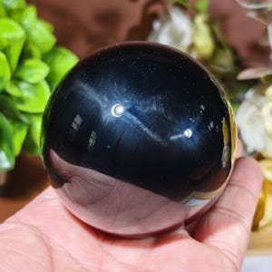 May include: A polished, spherical black stone with a smooth, reflective surface. The dark, opaque orb features subtle, shimmering inclusions that catch the light, creating a deep, lustrous appearance across its perfectly rounded, solid form.