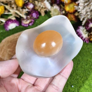 Può includere: Una pietra di selenite bianca traslucida e lucidata a forma di uovo al tegamino, caratterizzata da un centro in calcite arancione brillante, liscio e arrotondato, che ricorda un tuorlo, su una base piatta, irregolare e bianco latte.