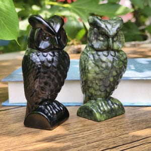 May include: Two owl figurines. One is a glossy black, the other is green with darker green marbling. Both are carved with detailed feather textures and have a stylized, upright posture. They are displayed on a wooden surface.