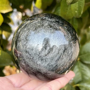 May include: A polished, spherical gemstone with a dark, iridescent sheen. The orb reflects light, showcasing swirling patterns of silver and gray. The stone is held in a hand, with green foliage in the background. A decorative crystal ball.