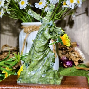 May include: A carved green stone sculpture depicting a figure kneeling while carrying a cross on their shoulder. The sculpture features a polished, mottled surface with varying shades of olive and forest green, set on a rectangular wooden base.