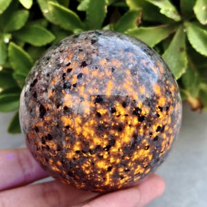 Puede incluir: Una esfera pulida con un patrón moteado de negro, gris y naranja. La esfera tiene una superficie lisa y reflectante y parece estar hecha de piedra. Las áreas naranjas brillan, creando un efecto visual llamativo. Es un objeto decorativo.