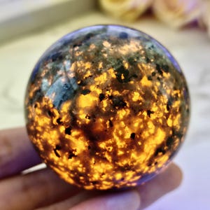 Peut inclure: Une sphère de pierre précieuse brillante avec un motif tacheté. L'orbite présente un mélange de tons sombres et terreux et d'inclusions jaune doré vif, créant un effet visuel captivant. La surface polie reflète la lumière, soulignant les détails complexes de la pierre. Environ 7 cm de diamètre.
