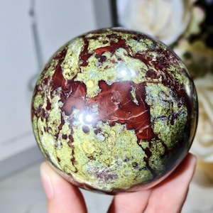 May include: A polished gemstone sphere, displaying a mottled pattern of deep red and olive green. The sphere is held in a hand, showcasing its smooth, rounded form. The stone's surface reflects light, highlighting its unique colour variations.