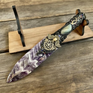 May include: An ornate dagger with a purple and white amethyst blade, set on a wooden stand. The handle features intricate metalwork, including a pentagram, bat wings, and colourful accents. The dagger is displayed on a wooden stand.