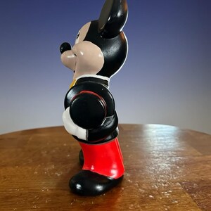 May include: A side view of a Mickey Mouse figurine. The figurine is wearing a black jacket with a red trim, red shorts, and black shoes. The figure has a black head and ears, a tan face, and a black nose.