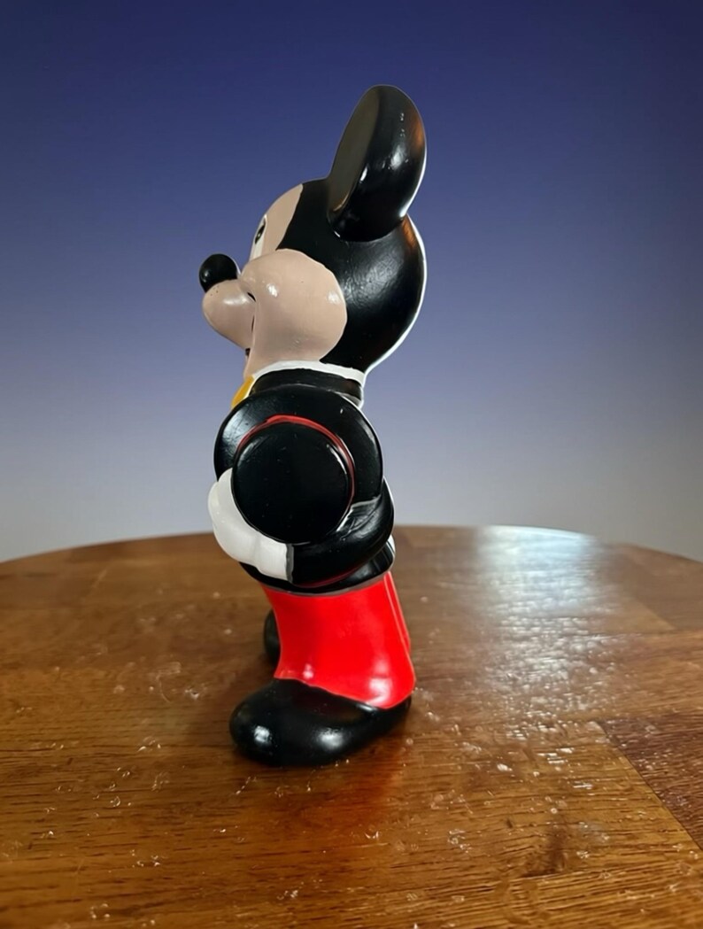 May include: A side view of a Mickey Mouse figurine. The figurine is wearing a black jacket with a red trim, red shorts, and black shoes. The figure has a black head and ears, a tan face, and a black nose.