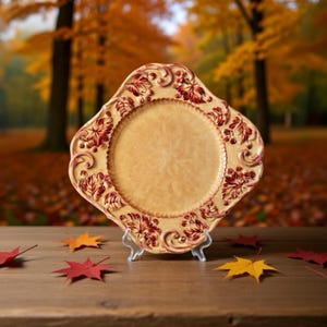 May include: A decorative, cream-colored plate with a raised, ornate design featuring burgundy-colored leaves and berries. The plate has a scalloped edge and is displayed on a clear stand, with autumn leaves scattered around it.