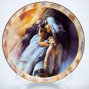 May include: Decorative plate featuring a Native American woman cradling a child. The artwork showcases soft colors, a gold rim, and a signature. The woman wears a feather in her hair, and the child is wrapped in a blue blanket.