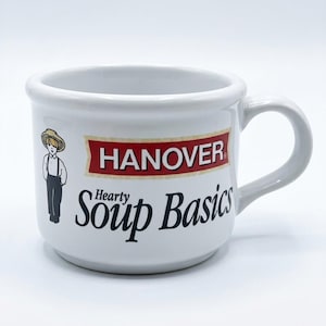May include: A white ceramic mug featuring the text Hanover in a red banner above the words Hearty Soup Basics in black lettering. To the left of the text is a small illustration of a person wearing a straw hat, white shirt, and dark trousers.