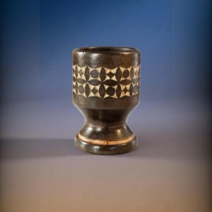May include: A dark brown wooden egg cup featuring a carved geometric pattern of intersecting circles and diamonds around the upper rim. The base is flared with a smooth, polished finish and a lighter wood accent ring near the bottom edge.