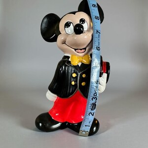 May include: A ceramic Mickey Mouse figurine, approximately 9 inches tall, dressed in a black tuxedo with a yellow bow tie and red shorts. The figurine has a smiling expression and is holding a small hat.