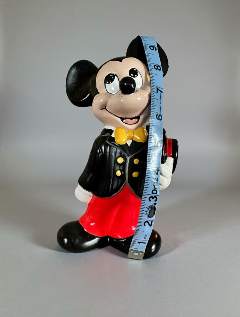 May include: A ceramic Mickey Mouse figurine, approximately 9 inches tall, dressed in a black tuxedo with a yellow bow tie and red shorts. The figurine has a smiling expression and is holding a small hat.