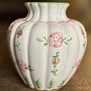 May include: White ceramic vase with a ribbed body and a flared top. The vase is adorned with pink roses and green leaves. The rim is green, and the vase has a delicate, hand-painted appearance.