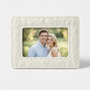 May include: A rectangular off-white picture frame featuring a raised, embossed floral pattern across the entire border. The frame holds a photograph of two individuals smiling together outdoors, with a soft-focus green background visible.