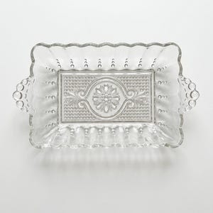 May include: A rectangular clear glass serving dish with a scalloped edge and two side handles. The base features a central embossed floral motif surrounded by a textured pattern of small raised dots, while the sides have vertical fluting.