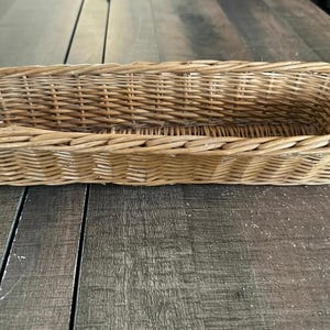 May include: A rectangular woven basket made of light brown natural wicker material. The basket features a long, narrow shape with a sturdy braided rim and a woven base, showcasing a consistent pattern of interlaced reeds throughout the structure.