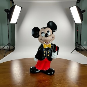 May include: A ceramic Mickey Mouse figurine, dressed in a black tuxedo with gold buttons, red pants, and black shoes. The figurine is holding a top hat and has a yellow bow tie. The background is a studio setup with lighting equipment.