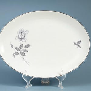 May include: An oval, white serving platter with a silver rim. The platter features a delicate black and white rose and leaf design. The platter is displayed on a clear stand against a blue background.