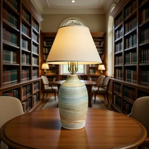 May include: A decorative table lamp with a cream-colored lampshade and a ceramic base. The base features a blue, green, and beige swirl pattern. The lamp is placed on a round wooden table in a library setting.