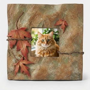 May include: A square picture frame with a textured, stone-like brown and green finish. The frame is decorated with three dimensional red maple leaves and thin, twisted metal wire accents. A photograph of a fluffy orange cat is inside the frame.