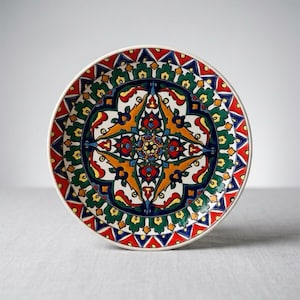 May include: A round ceramic plate featuring a vibrant, symmetrical pattern with intricate floral and geometric motifs. The design incorporates bold colors including deep blue, bright red, forest green, and golden yellow against a white background.