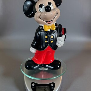 May include: A ceramic Mickey Mouse figurine in a tuxedo, red pants, and a yellow bow tie. The black and white figure is holding a top hat. The figurine is standing on a glass surface.