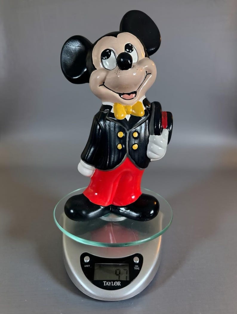 May include: A ceramic Mickey Mouse figurine in a tuxedo, red pants, and a yellow bow tie. The black and white figure is holding a top hat. The figurine is standing on a glass surface.