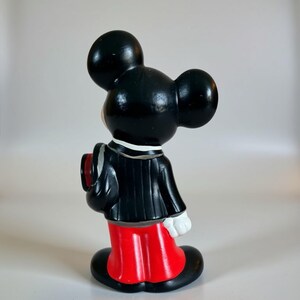 May include: A vintage Mickey Mouse figurine, viewed from the side. The figure is dressed in a black tuxedo with a red jacket and black shoes. The figurine has large black ears and white gloves, a classic Disney collectible.