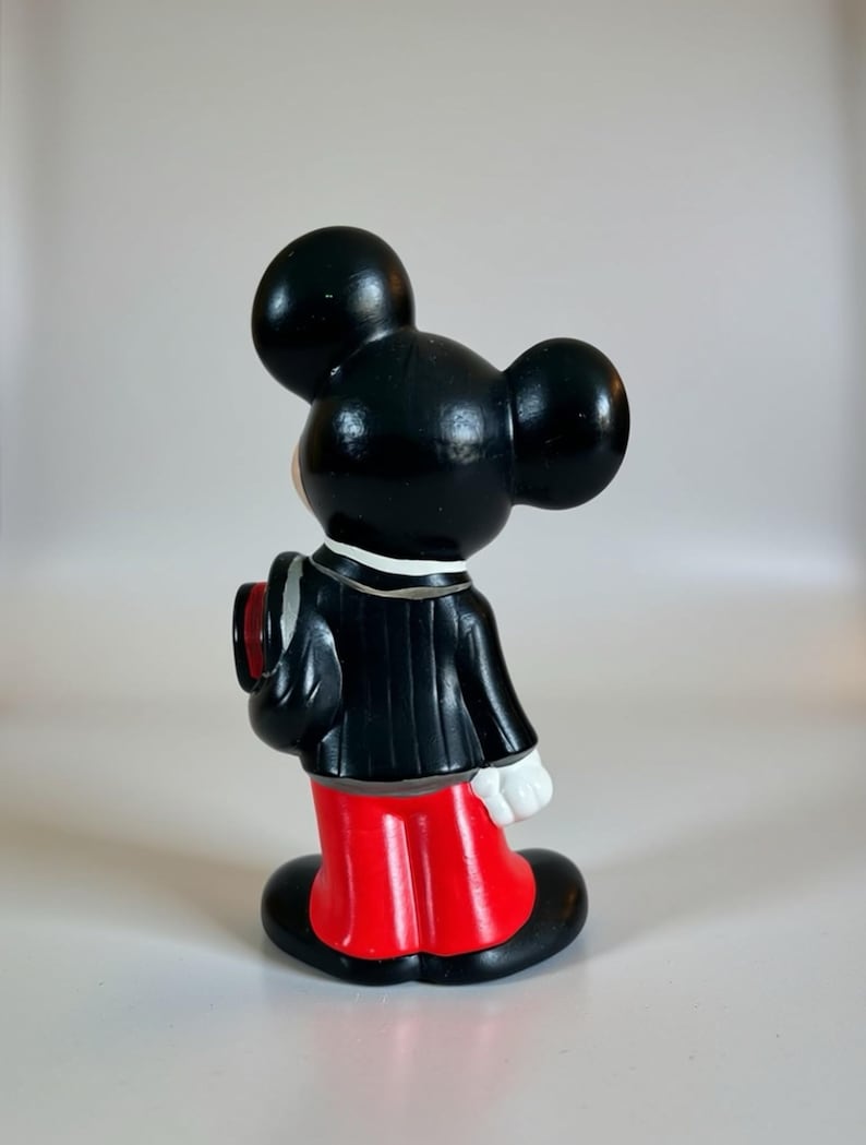 May include: A vintage Mickey Mouse figurine, viewed from the side. The figure is dressed in a black tuxedo with a red jacket and black shoes. The figurine has large black ears and white gloves, a classic Disney collectible.
