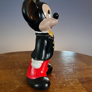 May include: A ceramic Mickey Mouse figurine in a classic outfit. The figure is black, red, and white, with a yellow bow tie. The figurine is standing on a wooden surface.