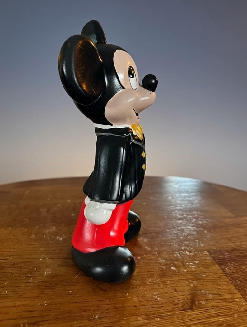 May include: A ceramic Mickey Mouse figurine in a classic outfit. The figure is black, red, and white, with a yellow bow tie. The figurine is standing on a wooden surface.