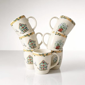 May include: A stack of six ceramic mugs. The mugs are off-white with a decorative band of yellow and green leaves around the top. Each mug features a birdhouse design with red hearts and a bird. Some mugs have a floral design.