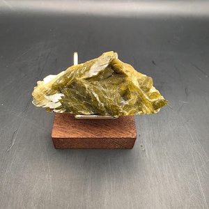 May include: A translucent, olive-green mineral specimen with a layered, crystalline structure. The mineral is mounted on a small, rectangular, dark brown wooden base. The background is a dark gray.