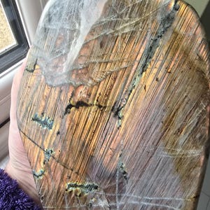 Puede incluir: Una losa de labradorita pulida y redondeada con un prominente brillo chatoyante dorado y bronce. La superficie muestra intrincadas estr&iacute;as lineales, fracturas internas naturales e inclusiones minerales oscuras en toda su cara.