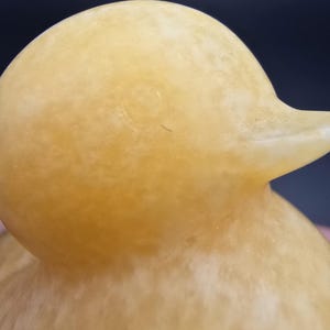 May include: Close-up of a yellow duck figurine. The duck has a smooth, matte finish and a rounded head with a small beak. The body has a textured appearance. The background is a dark blue and pink.