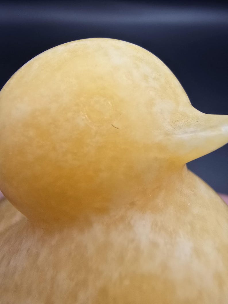 May include: Close-up of a yellow duck figurine. The duck has a smooth, matte finish and a rounded head with a small beak. The body has a textured appearance. The background is a dark blue and pink.