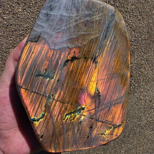 Puede incluir: Una piedra de labradorita pulida que presenta un despliegue vibrante de destellos iridiscentes en naranja, amarillo y rosa sobre su superficie. La piedra tiene una forma lisa y redondeada con inclusiones minerales oscuras y estr&iacute;as finas.