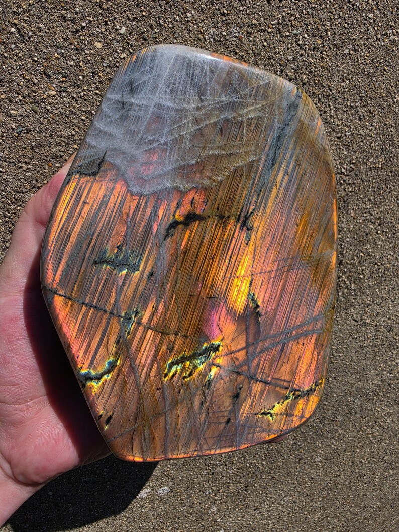 Puede incluir: Una piedra de labradorita pulida que presenta un despliegue vibrante de destellos iridiscentes en naranja, amarillo y rosa sobre su superficie. La piedra tiene una forma lisa y redondeada con inclusiones minerales oscuras y estr&iacute;as finas.