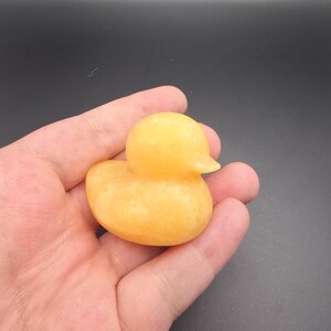 May include: A small, yellow duck figurine with a smooth, polished surface. The duck has a rounded body, a small beak, and a simple, classic design. The figurine is held in a hand against a dark background.