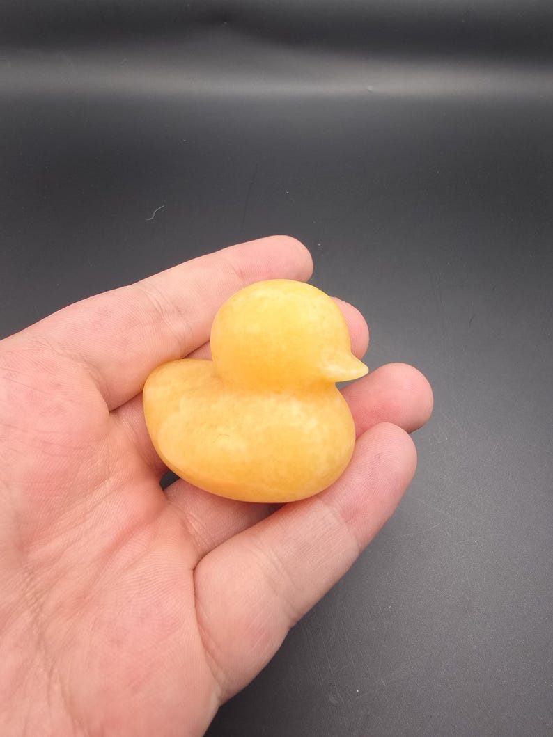 May include: A small, yellow duck figurine with a smooth, polished surface. The duck has a rounded body, a small beak, and a simple, classic design. The figurine is held in a hand against a dark background.