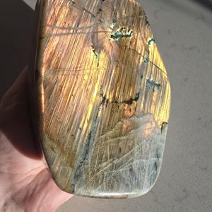 Puede incluir: Una losa de piedra labradorita pulida con una forma rectangular redondeada y lisa. La superficie muestra una vibrante iridiscencia dorada y bronce con estr&iacute;as lineales visibles e inclusiones minerales oscuras y dentadas.